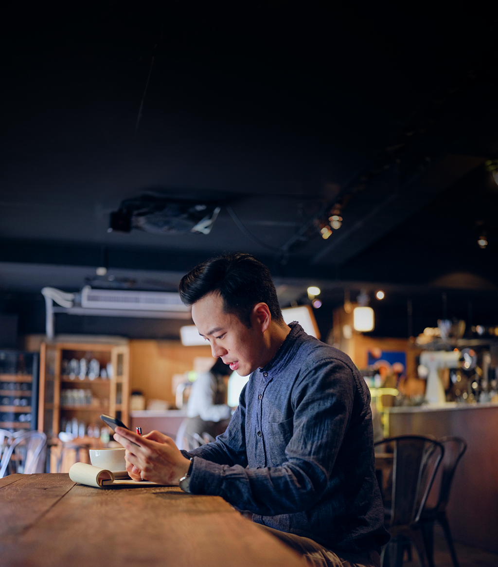 Asian man in coffee sitting at large table with notepad and coffee mug looking at his cell phone.