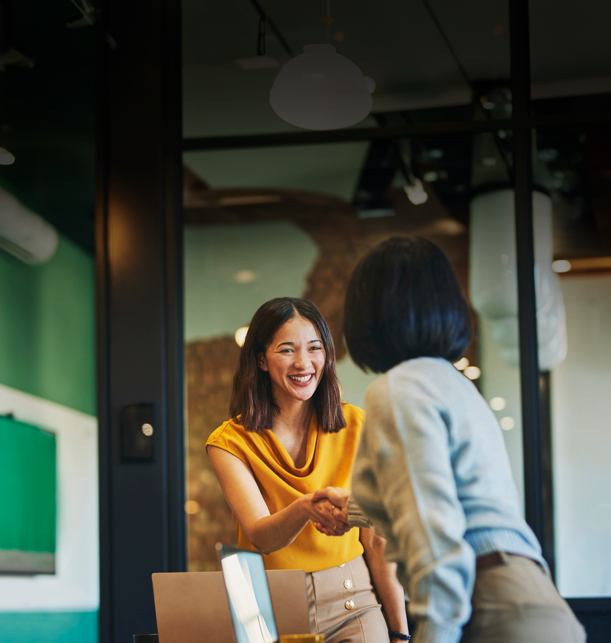 young smiling woman in professional clothing standing and shaking hands with another woman in conference room 