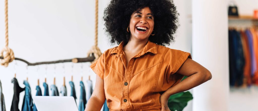 Black female small business owner smiling cheerfully while standing in her shop. Happy businesswoman managing her clothing orders on a laptop. Black female entrepreneur running an online clothing store.